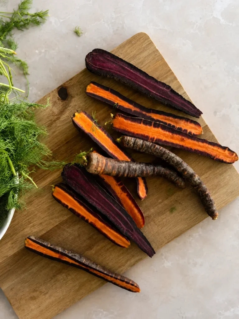 Purple carrots sliced lengthwise on a wooden cutting board, showing vivid orange and deep violet interiors on a pale stone surface.