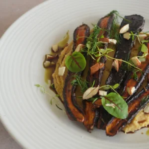 Vegan roasted purple carrots plated over creamy chickpea purée and finished with herbs, microgreens, chopped almonds, olive oil and glaze on a white ceramic plate.