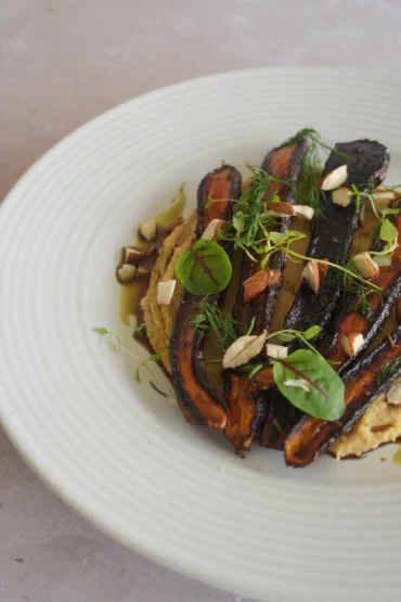 Vegan roasted purple carrots plated over creamy chickpea purée and finished with herbs, microgreens, chopped almonds, olive oil and glaze on a white ceramic plate.