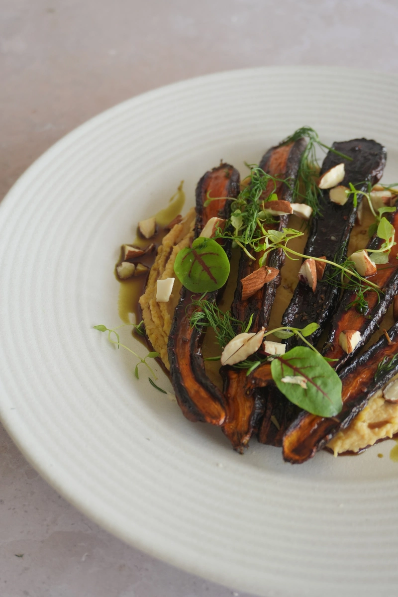 Vegan roasted purple carrots plated over creamy chickpea purée and finished with herbs, microgreens, chopped almonds, olive oil and glaze on a white ceramic plate.
