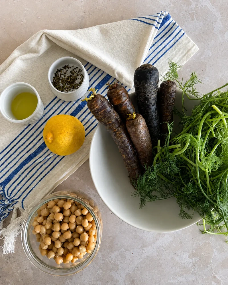 Vegan roasted heirloom carrots ingredients with purple carrots, chickpeas, lemon, olive oil and fresh fennel fronds arranged on a pale stone surface.