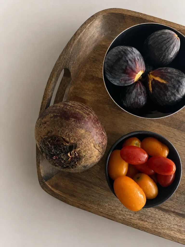 Overhead photo of a wooden tray holding a whole beetroot, a black bowl of fresh dark figs, and a small bowl of red and yellow cherry tomatoes on a light neutral background.