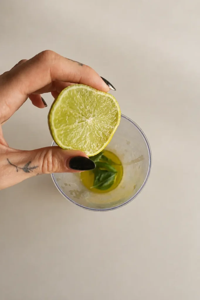 Hand holding half a lime above a small cup with olive oil and basil leaves, preparing the basil dressing for the fig carpaccio.