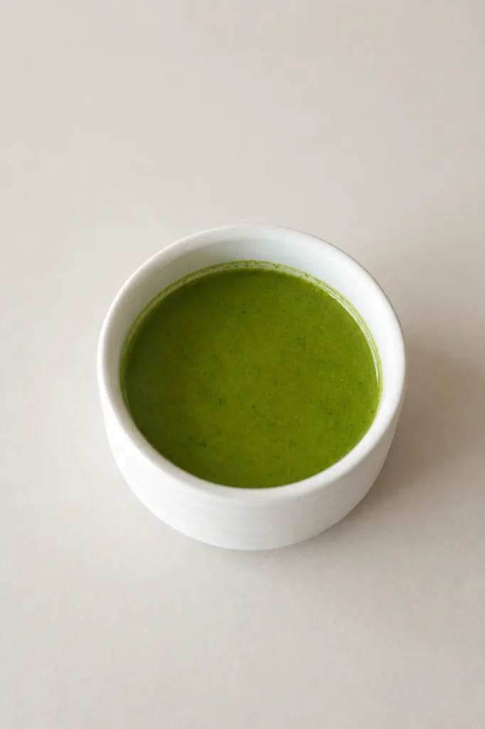 Small white bowl filled with bright green basil oil on a light neutral background.