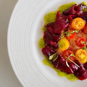 Close-up of fig carpaccio plated with deep red beetroot ribbons, yellow and red cherry tomatoes, basil oil, and microgreens on a white ridged plate.