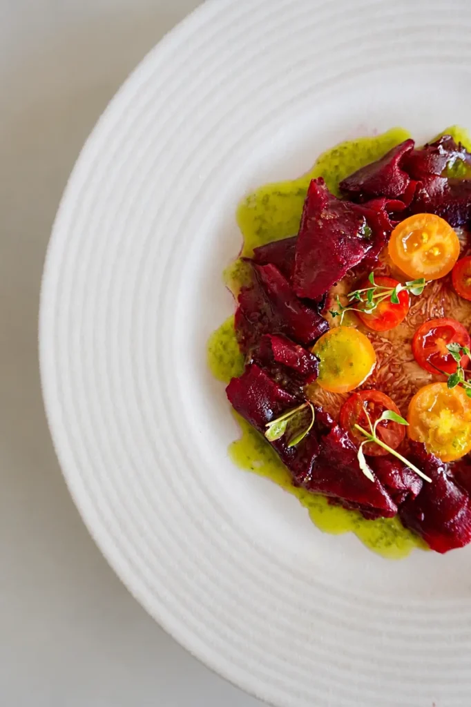 Close-up of fig carpaccio plated with deep red beetroot ribbons, yellow and red cherry tomatoes, basil oil, and microgreens on a white ridged plate.
