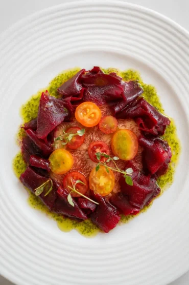 Overhead view of fig carpaccio topped with deep red beetroot ribbons, yellow and red cherry tomatoes, microgreens, and bright basil oil on a white ridged plate.