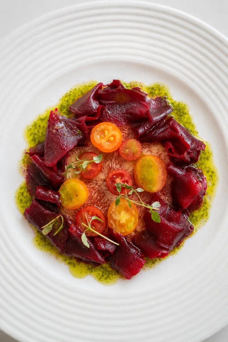 Overhead view of fig carpaccio topped with deep red beetroot ribbons, yellow and red cherry tomatoes, microgreens, and bright basil oil on a white ridged plate.