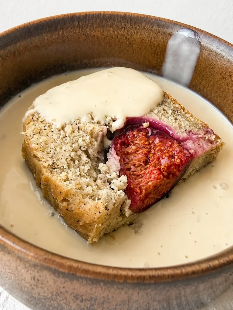 A slice of fig bread served in a rustic brown bowl with vegan crème anglaise, showing a soft crumb and a baked fig inside.