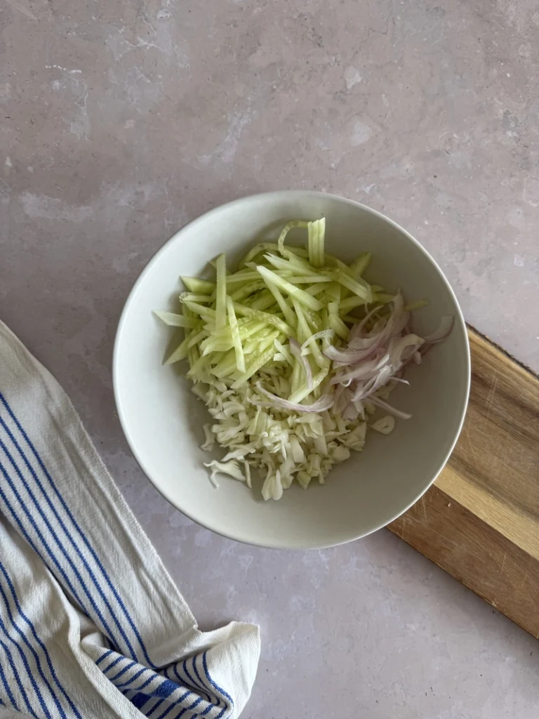 Kachumber salad preparation with julienned cucumber, shredded cabbage and thinly sliced red onion in a bowl.
