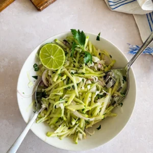 Kachumber salad in a white bowl with julienned green apple, cabbage, red onion, fresh herbs and a lime half, served with two spoons on a light stone surface.