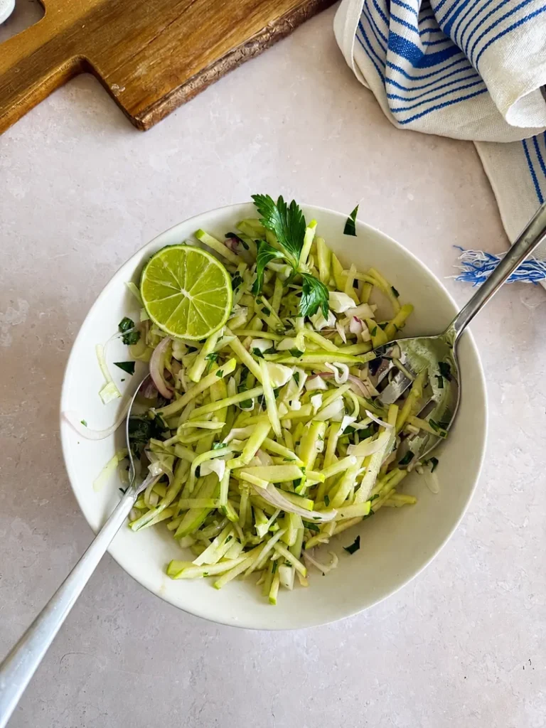 Kachumber salad in a white bowl with julienned green apple, cabbage, red onion, fresh herbs and a lime half, served with two spoons on a light stone surface.