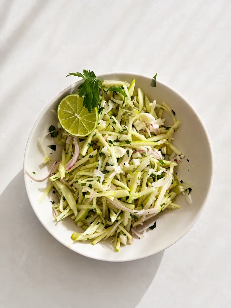 Kachumber salad with julienned cucumber, green apple, cabbage, red onion, fresh herbs and lime, served in a white bowl on a light stone surface.