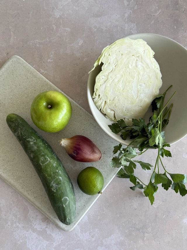 Fresh cabbage, cucumber, green apple, shallot, lime and parsley arranged on a stone surface in soft natural light.