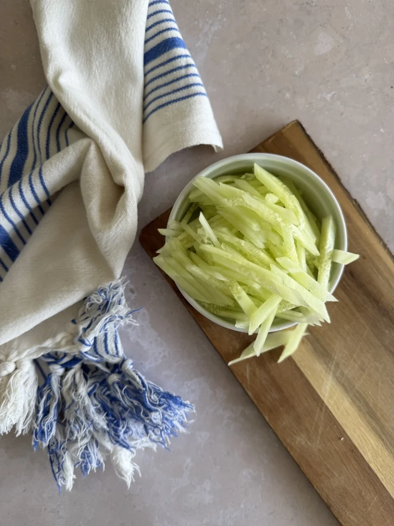 Julienned cucumber arranged on a wooden cutting board beside a blue-and-white striped kitchen towel on a light stone surface.