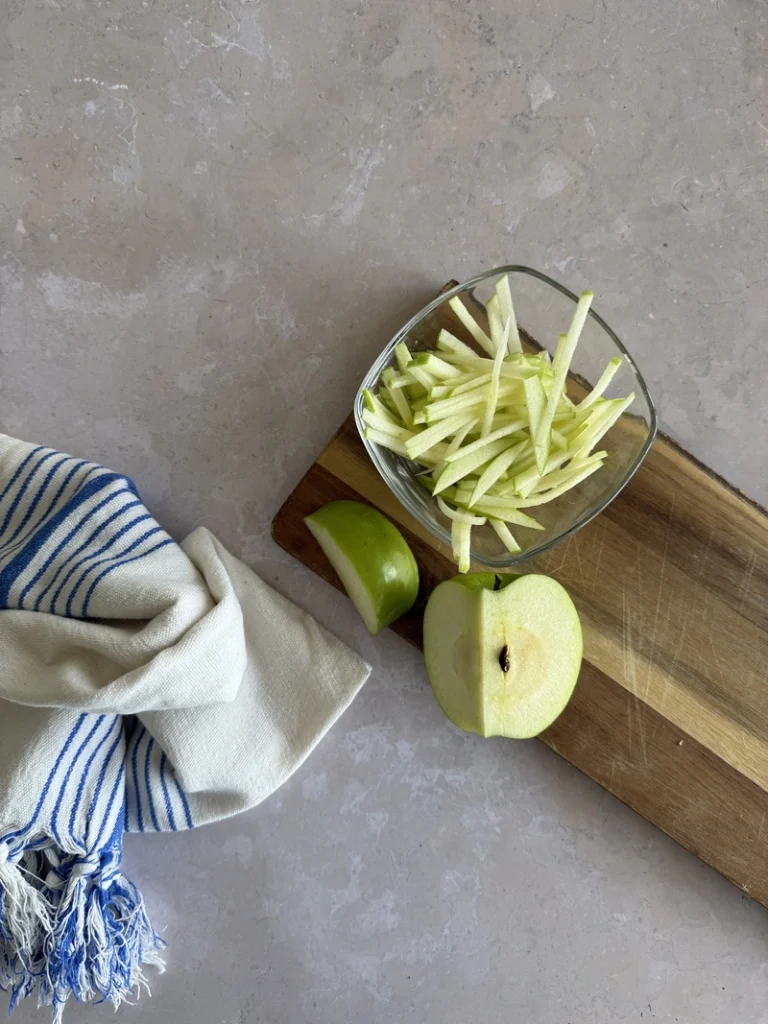 Julienned green apple on a wooden cutting board, prepared as an ingredient for kachumber salad, beside a blue-and-white striped kitchen towel.