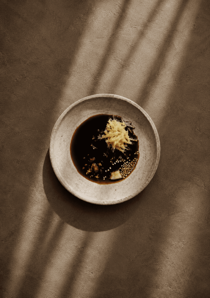 Tamari marinade on a ceramic plate with grated ginger, sesame seeds and a little dried chilli, styled on a warm textured background with soft natural shadows.