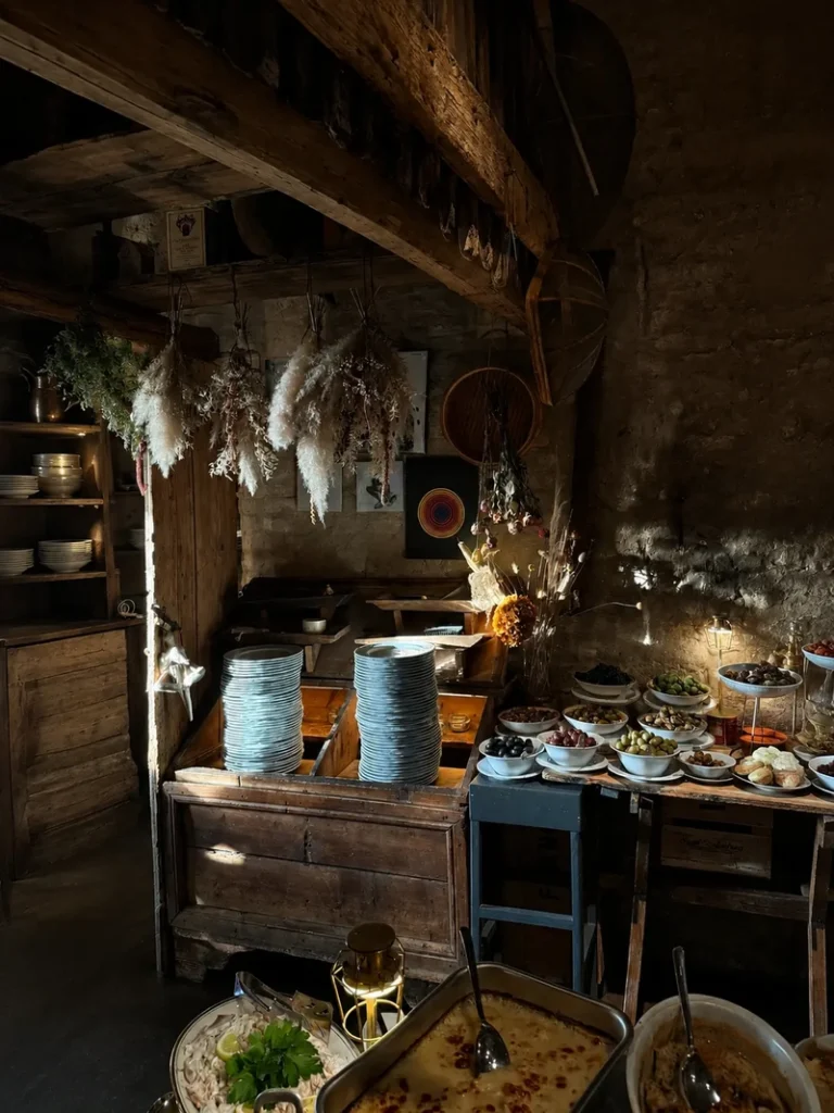 Rustic buffet corner with stacked plates, hanging dried flowers, bowls of food and warm light inside a cozy countryside restaurant.