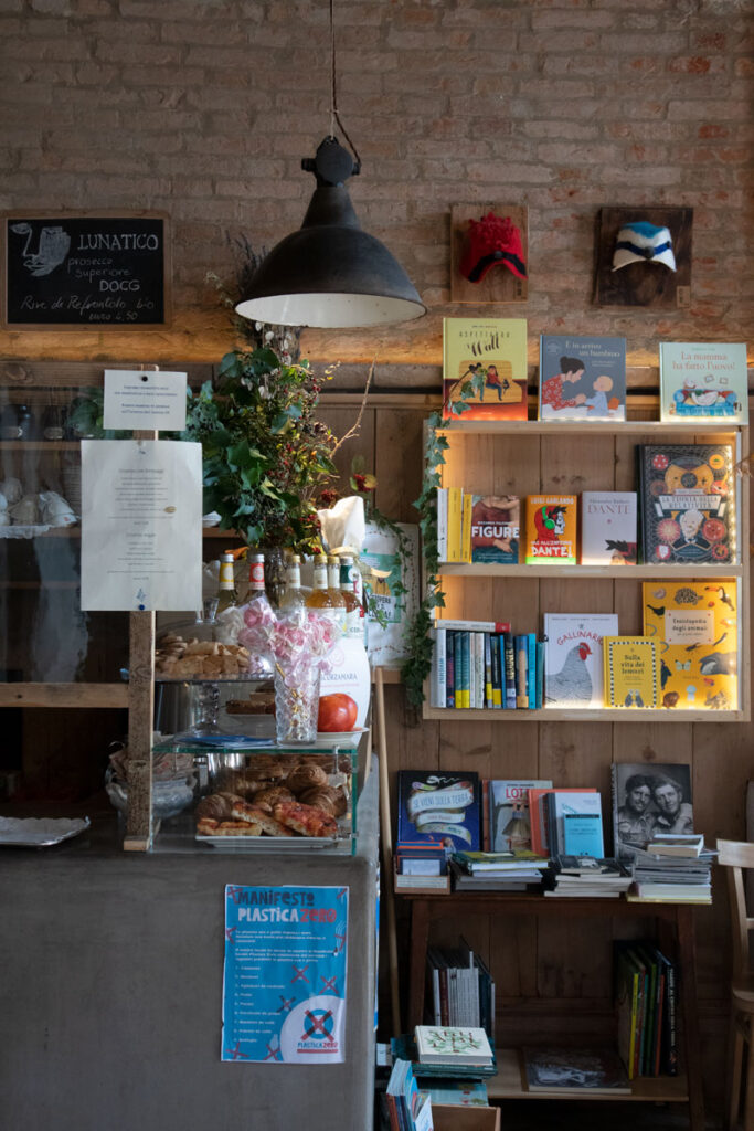 Interior of Sulla Luna Venezia, a vegan-friendly bookshop bistro in Venice, Italy, with bookshelves, pastries and rustic brick walls.