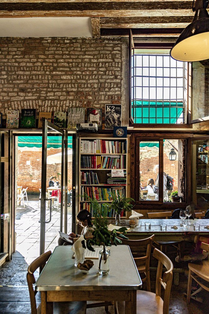 Dining area at Sulla Luna Venezia in Venice, Italy, with wooden tables, bookshelves, exposed brick walls and an open view onto the courtyard.