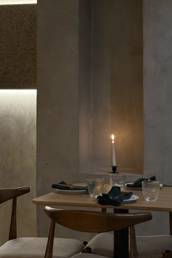 Dimly lit minimalist restaurant table set with white plates, dark napkins and clear glasses, illuminated by a single candle against warm textured walls and wooden chairs.