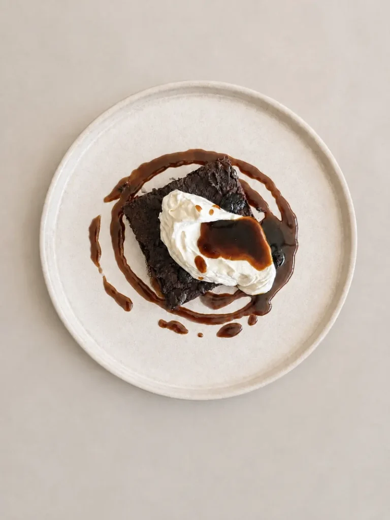High-protein vegan bean brownie served with whipped coconut cream, dark chocolate shards and miso caramel on a light ceramic plate.