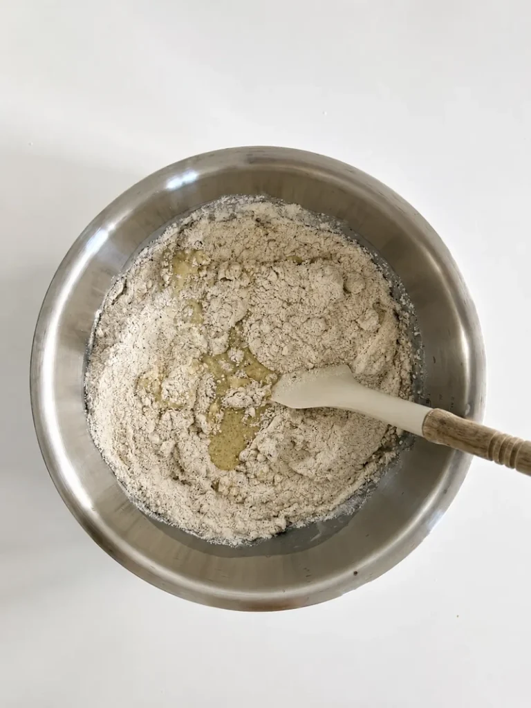 Overhead view of the gluten-free flour mixture being added to the wet ingredients for vegan fig bread with fresh figs, shown in a stainless-steel bowl with a spatula.
