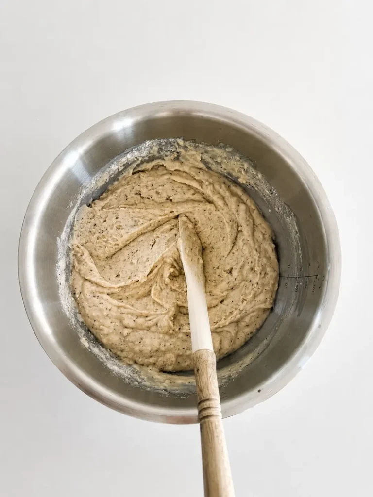 Overhead view of the thick gluten-free batter for vegan fig bread with fresh figs after mixing, in a stainless-steel bowl with a spatula and soft neutral natural light.