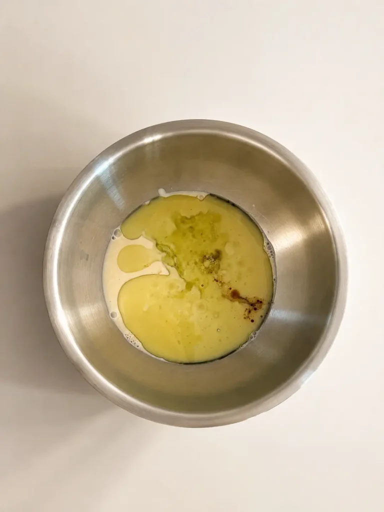 Overhead view of the wet ingredients for vegan fig bread with fresh figs in a stainless-steel mixing bowl, including soy yogurt, oil, and vanilla, lit with soft neutral natural light.