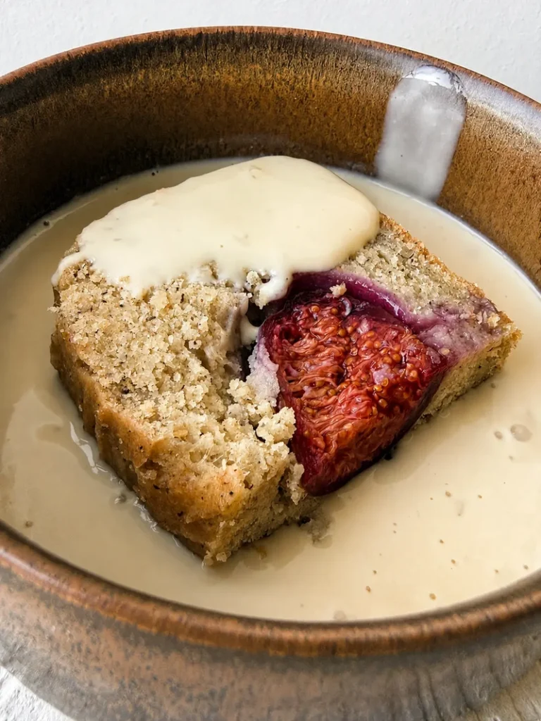 Close-up of vegan gluten-free fig bread with fresh figs served in a bowl with silky vegan crème anglaise, showing the whole fig baked into the centre of the slice.