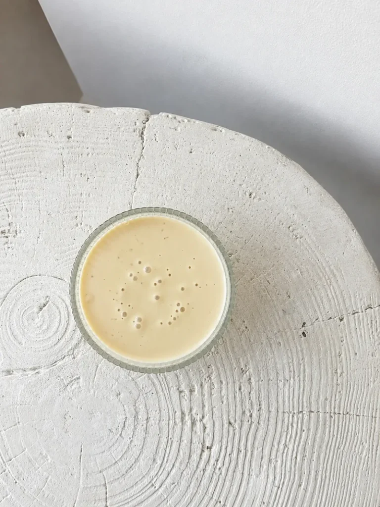 Overhead view of vegan crème anglaise in a small glass bowl, showing its smooth, creamy texture on a textured white surface with soft natural light.