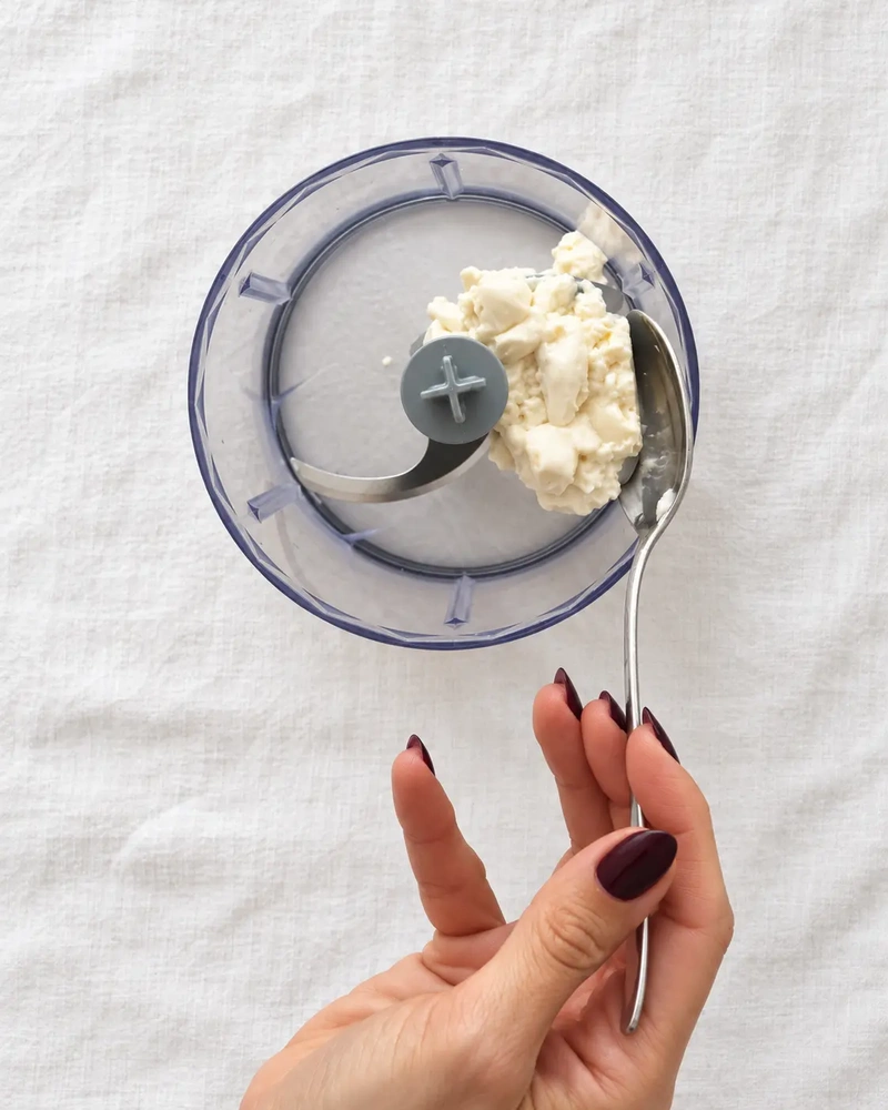 Silken tofu being added with a spoon to a small food processor on white linen for tofu chocolate pudding.