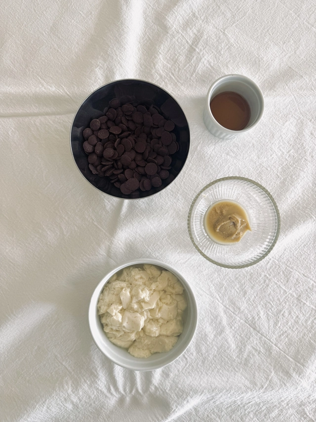 Ingredients for tofu chocolate pudding arranged on white linen, including silken tofu, dark chocolate, tahini and unsweetened soy milk.