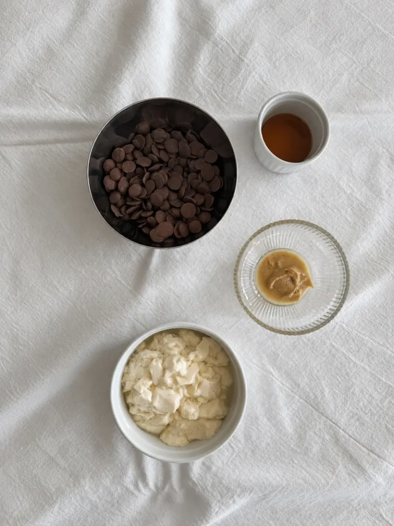 Ingredients for chocolate tofu mousse arranged on a white cloth, including silken tofu, dark chocolate, tahini, and maple syrup.
