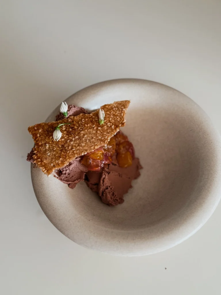 Chocolate tofu mousse plated in a beige bowl with macerated fruit, a sesame tuile, and small white edible flowers, photographed from above.