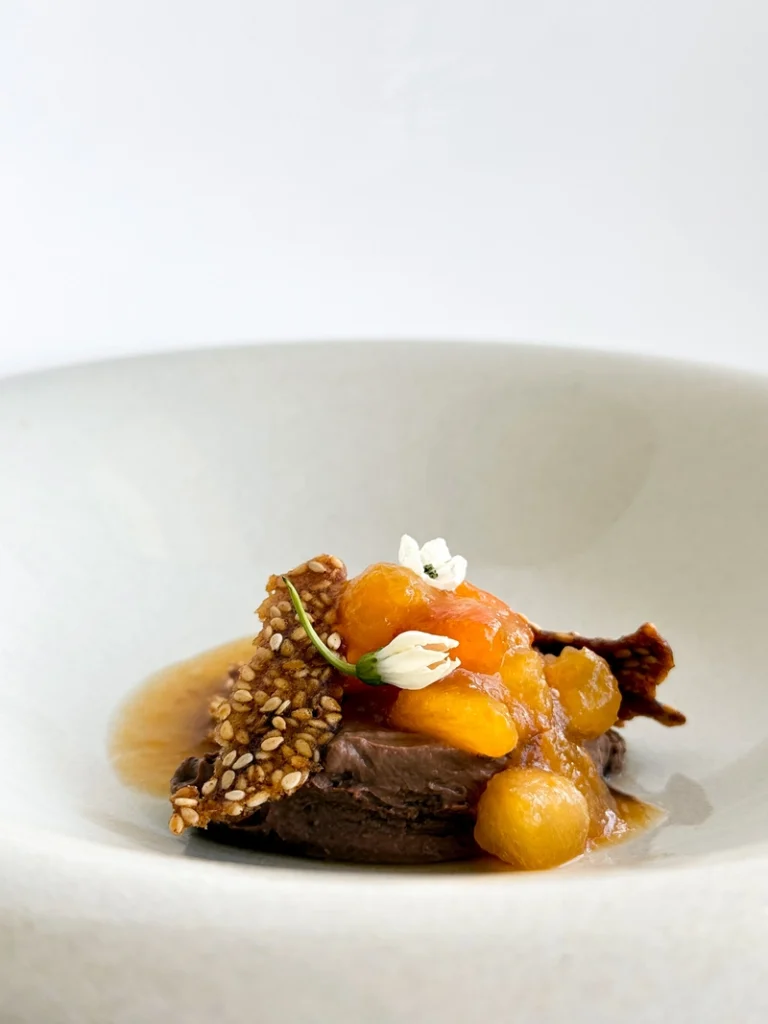 Silken tofu chocolate mousse topped with macerated plums, a crisp sesame tuile, and small white edible flowers.