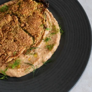 Vegan eggplant schnitzel with a crispy golden coating, served over creamy white bean and roasted tomato hummus and finished with fresh herbs and olive oil.