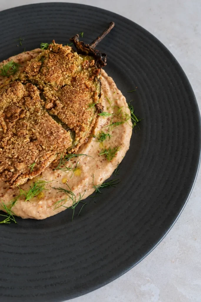 Vegan eggplant schnitzel with a crispy golden coating, served over creamy white bean and roasted tomato hummus and finished with fresh herbs and olive oil.