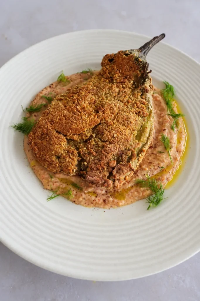 Vegan eggplant schnitzel with a crisp golden coating, served over creamy white bean and roasted tomato hummus with olive oil and fresh dill.