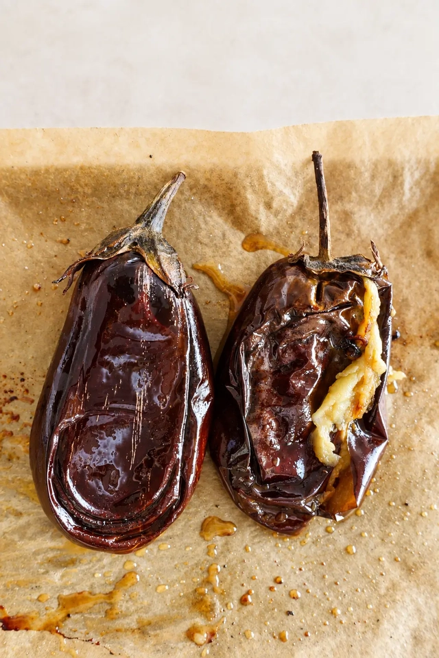 Two whole roasted eggplants on parchment paper, softened and wrinkled after baking, ready to be flattened and breaded for vegan eggplant schnitzel.