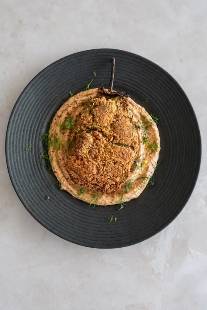 Vegan eggplant schnitzel with a crisp golden coating, served over creamy white bean and roasted tomato hummus with olive oil and fresh dill.