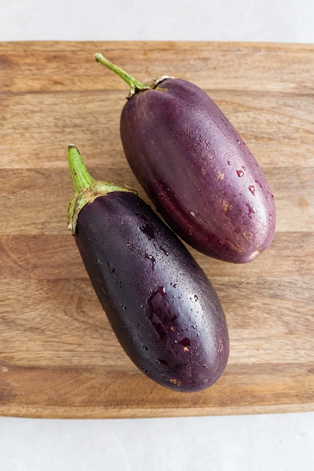 Two fresh whole eggplants on a wooden board, photographed from above in soft natural light, ready to be baked for vegan eggplant schnitzel.