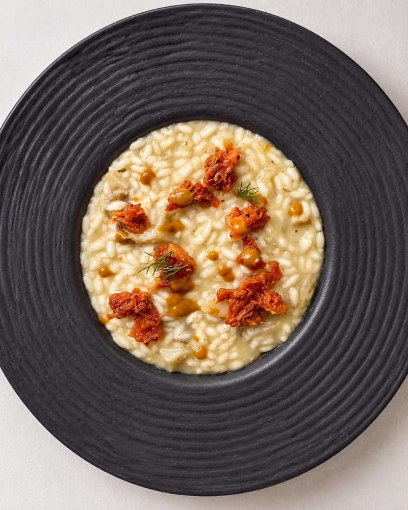 Creamy vegan risotto served in a black textured bowl, topped with sun dried tomatoes, herbs and a savoury sauce.