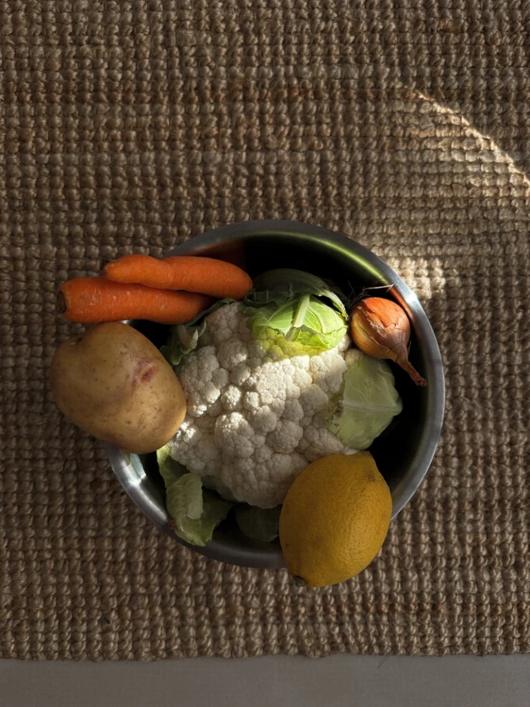 Ingredients for lemon carrot cauliflower soup, with cauliflower, carrots, potato, onion and lemon arranged in a metal bowl on a textured natural fiber background.