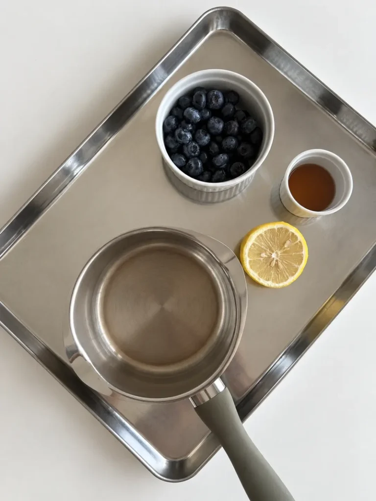 Ingredients for vegan panna cotta with blueberry coulis, including fresh blueberries, maple syrup, lemon, and a small saucepan arranged on a stainless steel tray.