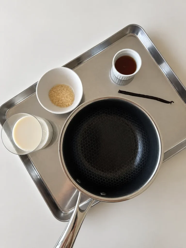 Ingredients for vegan panna cotta, including soy milk, agar agar flakes, maple syrup, vanilla, and a saucepan arranged on a stainless steel tray.