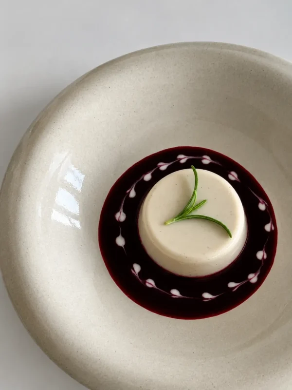 Vegan panna cotta with blueberry coulis, decorated with delicate yogurt drops and a fresh rosemary sprig on a large ceramic plate.
