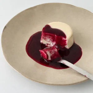 Vegan panna cotta with blueberry coulis served on a ceramic plate, with one bite cut out and a spoon holding a piece of the panna cotta covered in sauce.