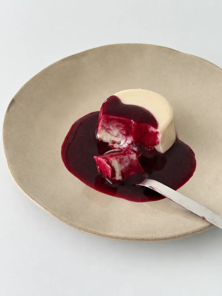 Vegan panna cotta with blueberry coulis served on a ceramic plate, with one bite cut out and a spoon holding a piece of the panna cotta covered in sauce.
