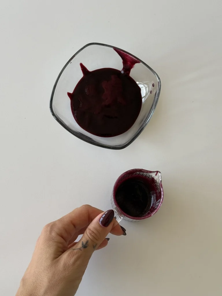 Blueberry coulis for vegan panna cotta being poured from a small glass jug beside a bowl of blended blueberries on a white surface.